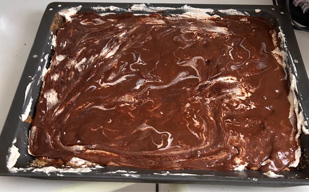 Schokoüberzogener Kuchen im Blech. Die Kuvertüre ist von weißen Sahnespuren durchzogen, am Backblech kleben auch Sahnereste.