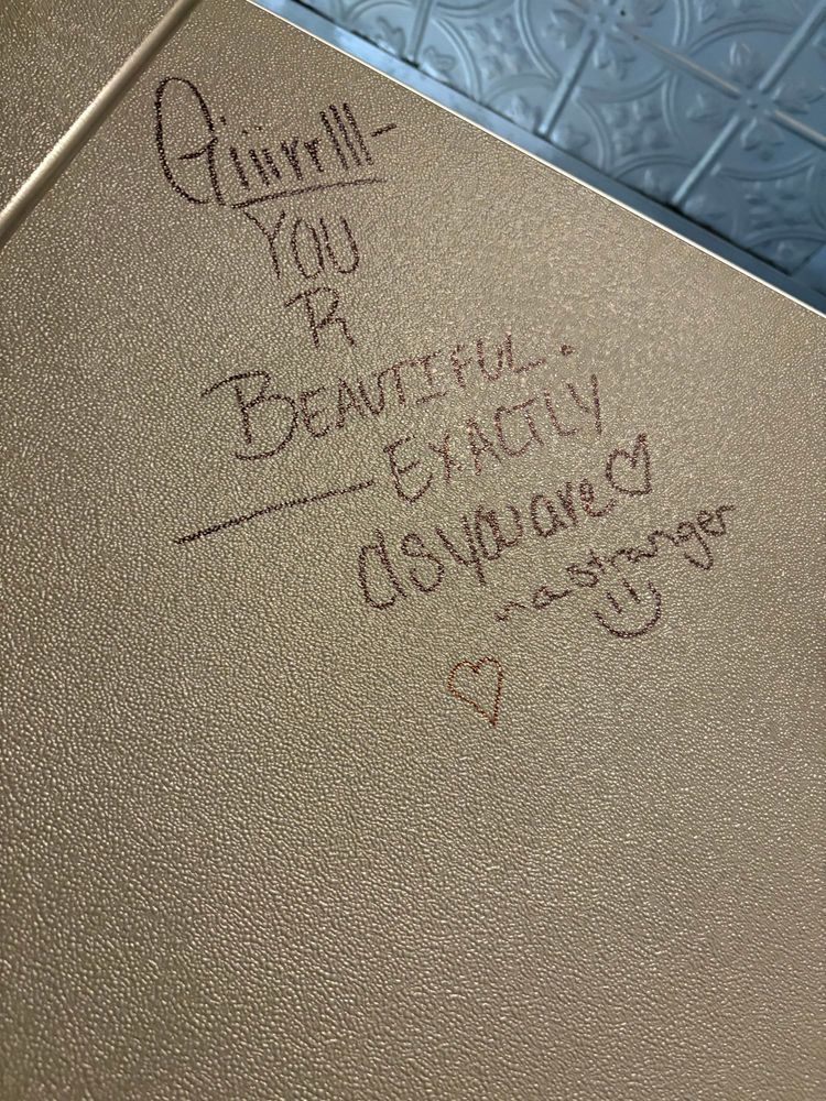 A wall in a public bathroom on which someone has written in black sharpie, “Girl, you are beautiful exactly as you are. A stranger.“