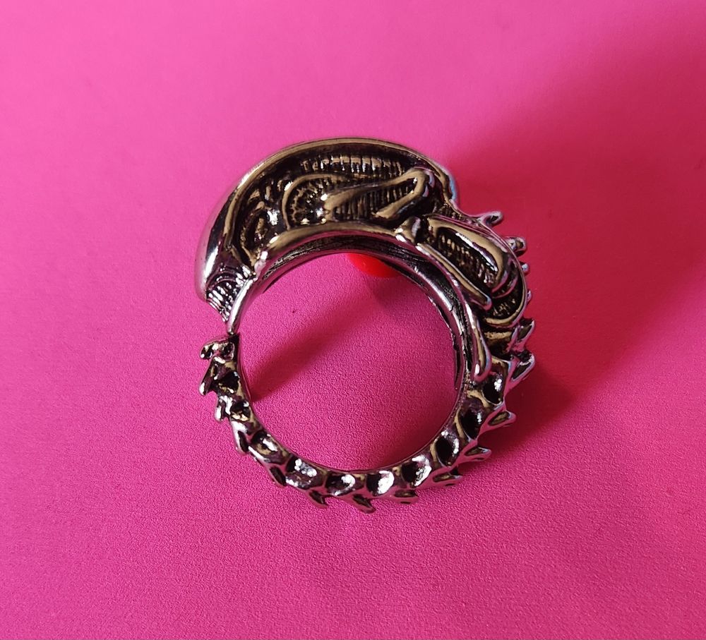 A silver ring on a pink background. The ring is of a Xenomorph head and body that wraps around to form the ring. The head of the Xenomorph is quite tall and the spine has segments that surprisingly doesn't hurt. 