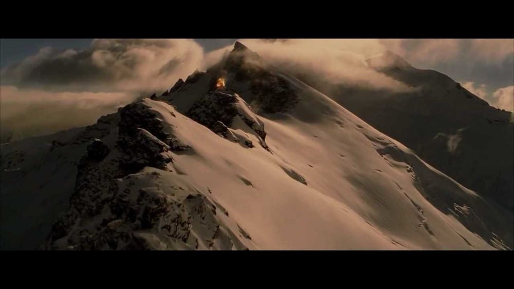 screenshot of the beacons of Gondor being lit in the Return of the King film.  A snow covered mountain in low yellow light—sunrise or sunset. There is a beacon of fire on the ridge.