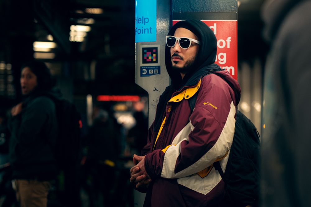 A young man in a hoodie wearing white sunglasses leans against a pole, hands clasped in front of him, in a subway station.