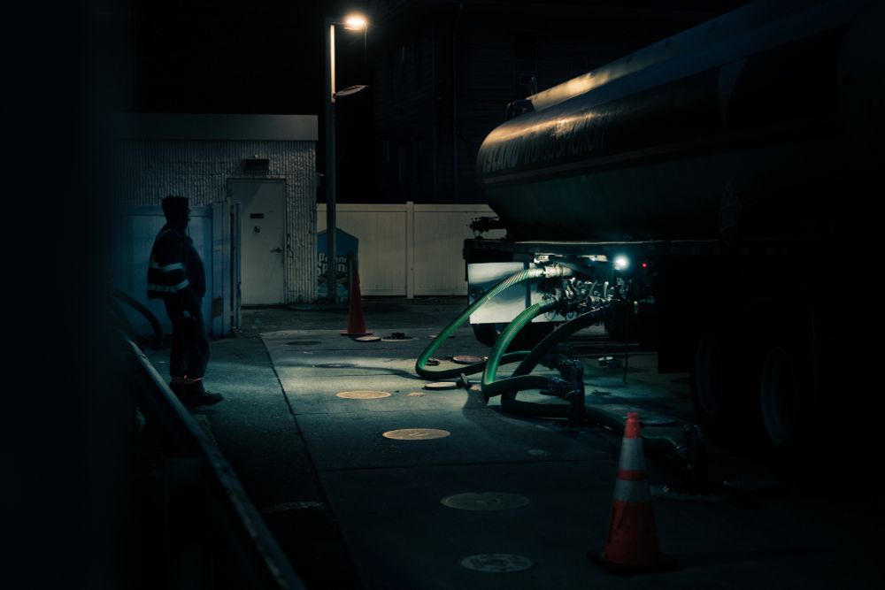 An starkly lit evening scene of a tanker truck refueling a gas station while the driver stands nearby observing. 