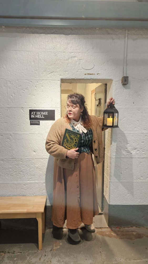 Same woman as in the first image, in full body holdling a lantern and a green book with a decorative colour in the doorway of a old Victorian time jail cell turned history museum with the placard At Home in Hell