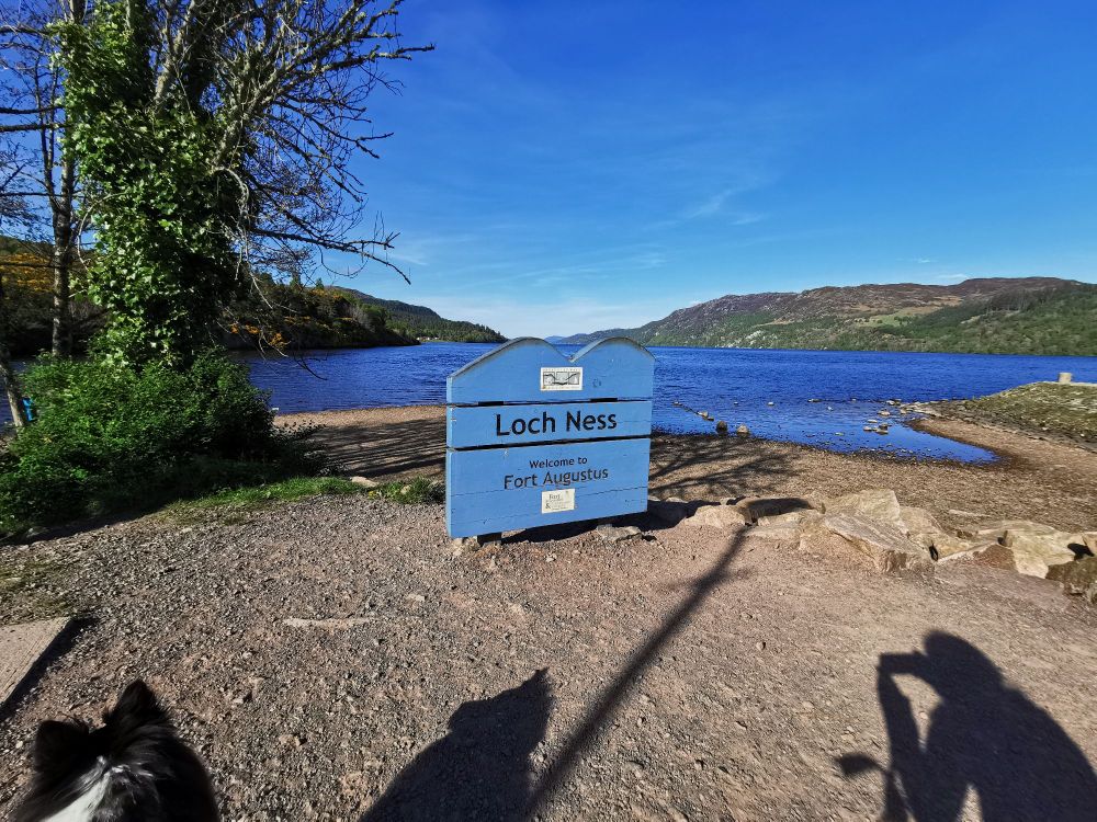 Sign with Loch Ness Welcome to Fort Augustus in front of Loch Ness and Mountains. On the left side is a dog. 