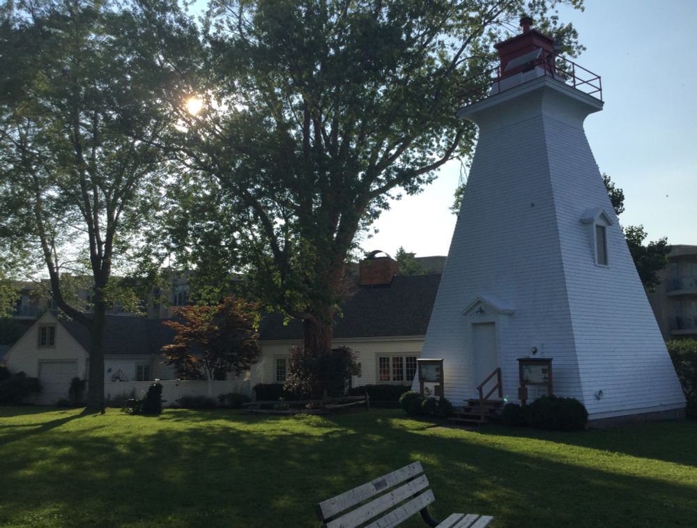 Photo taken in Niagara-on-the-Lake of a white lighthouse next to a couple of thick-trunked trees on a green lawn. 