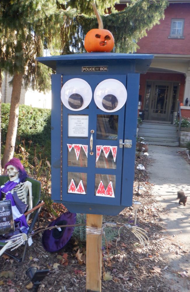 A little free library that usually looks like a TARDIS has been dressed up with googly eyes and bloody fangs. A small jack-o'-lantern sits on top of it.  Bonus black squirrel on the sidewalk next to it.  