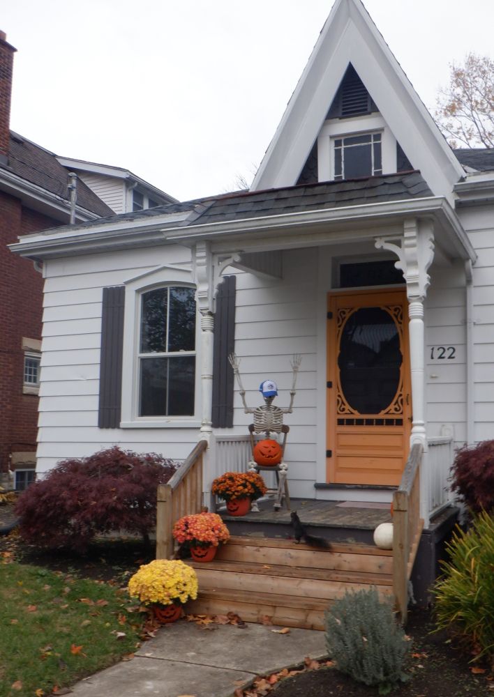 Photo of a skeleton sitting on a front porch, wearing a Blue Jays baseball cap. Bonus black squirrel on the porch steps.