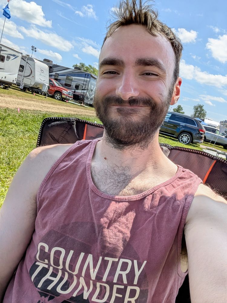 Sitting in a camping chair with a faded red/maroon tank top. A big smile on my face. Hair is spiked up in front a little bit