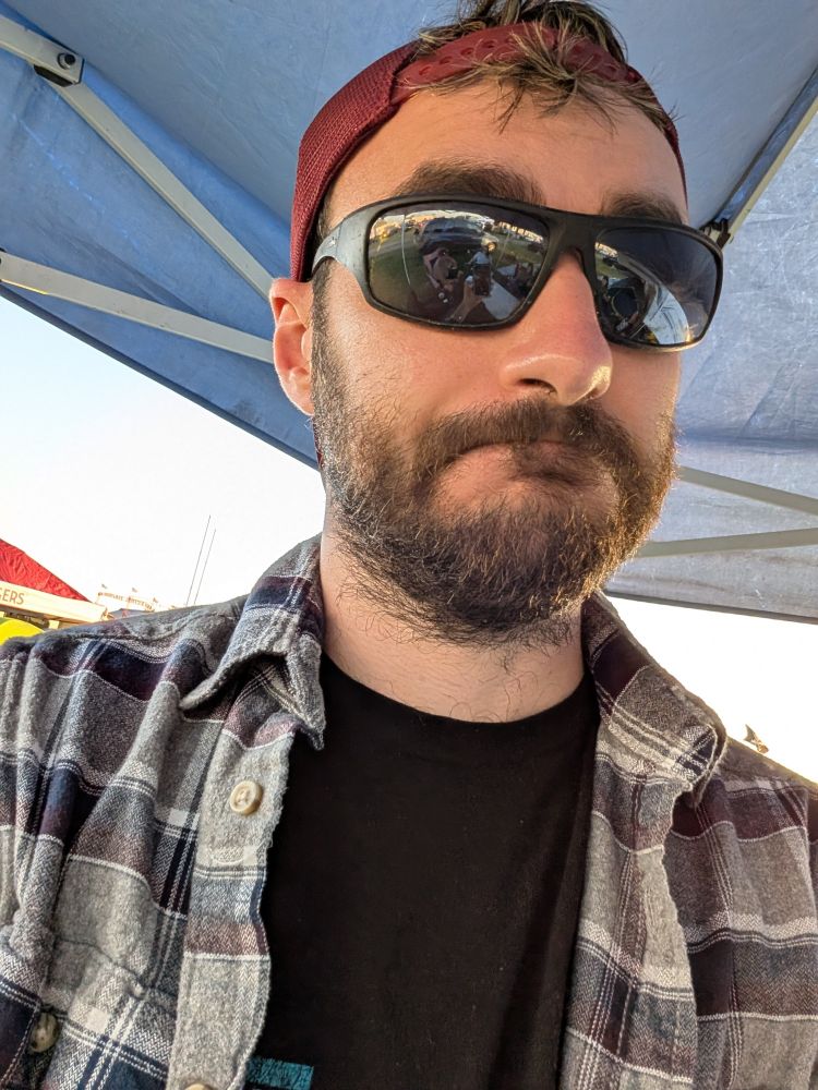 Selfie wearing a flannel with sunglass and backwards maroon hat. Possibly a "bear face" smile (close but not quite)