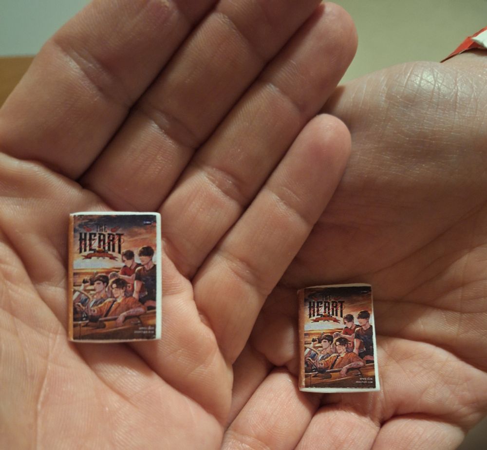 Two open palms are held together to display two miniature books. Each tiny book fits easily within the hands, emphasizing their small size. The covers of both books are identical, featuring an illustrated group of four young men sitting together in a sunset-lit scene with the title “THE HEART KILLERS” in bold stylized letters at the top. The artwork has a vintage or cinematic poster feel, and the young men appear to be part of a team, possibly referencing a show or story. 
