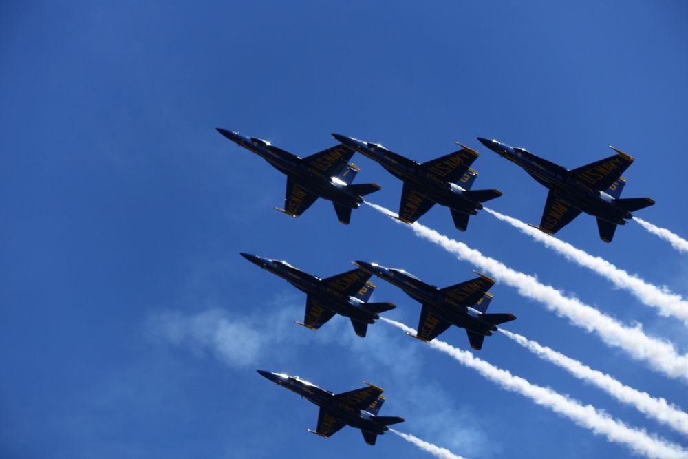 A photo of the Blue Angels flying in formation, trailing smoke in a blue sky at relatively close distance.