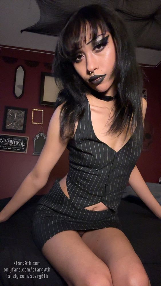 Star sitting on bed looking at the camera wearing a pinstriped vest and matching skirt 