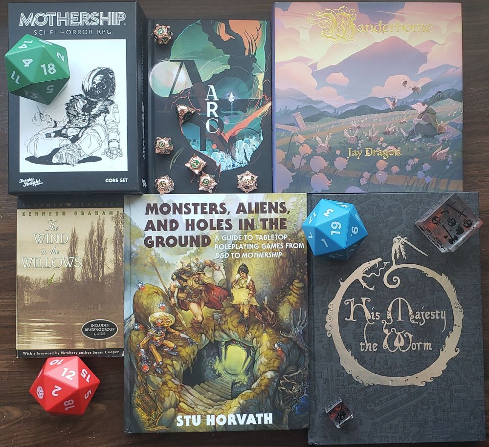 An overhead photograph of a table with ttrpg books and dice on it. The books include from left to right top to bottom: "Mothership" TTRPG box set, "ARC", "Wanderhome", "The Wind in the Willows", "Monsters, Aliens, and aholes in the Ground" by Stu Horvath, and "His Majesty the Worm". Atop and between the books are a various polyhedral dice. 1 set of 7, 3 large foam dice, and 2d6 gelatinous cube dice with skulls for pips
