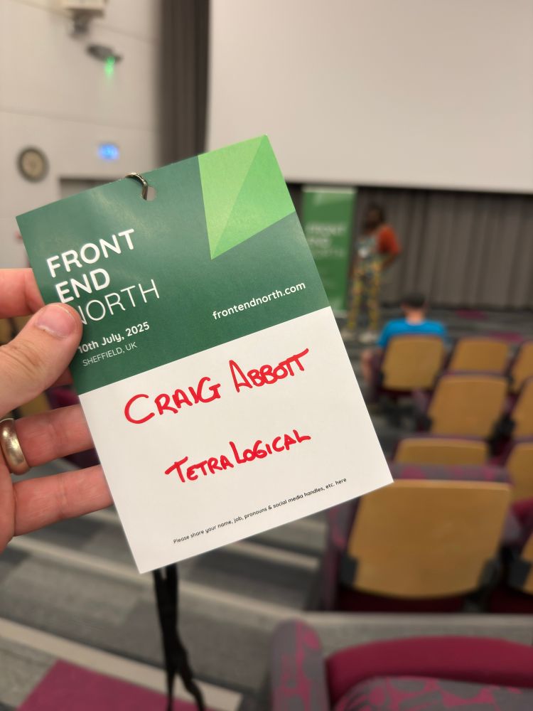 A lanyard and name card being held up in front of a projector screen in a lecture hall, it reads Front End North, Craig Abbott, TetraLogical.