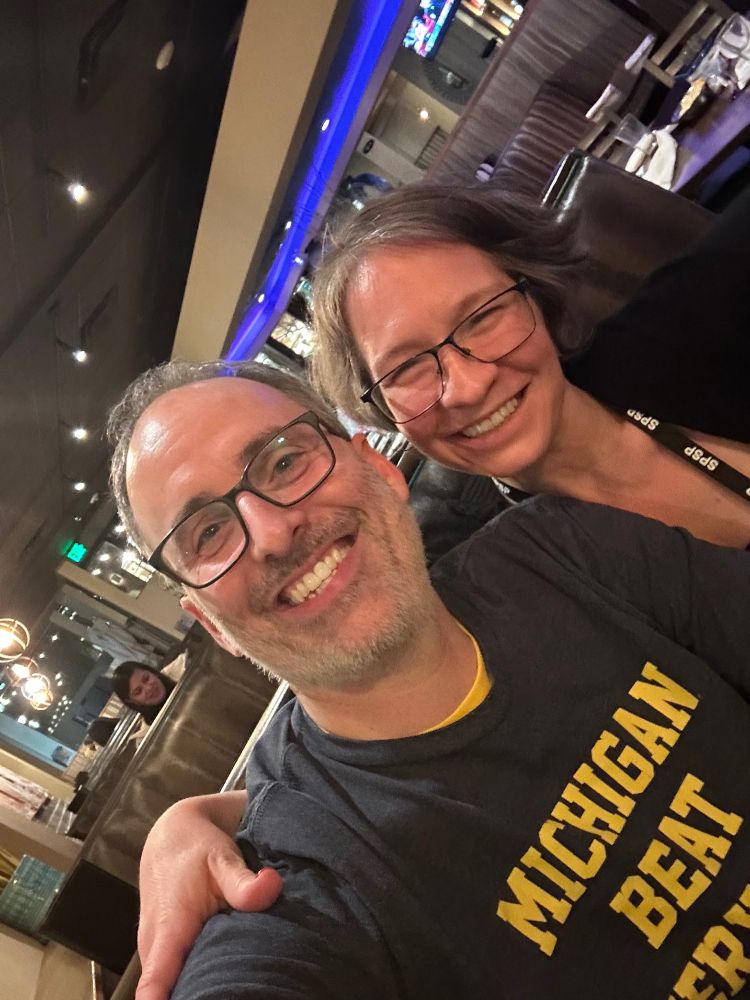 An image of Sam Sommers and Liz Page-Gould in Denver, Colorado, US in February 2025. Sam is taking the picture as a selfie and thus has his arm outstretched toward the camera. Liz has her arm around Sam's shoulder. Both people are smiling and looking at the camera.
