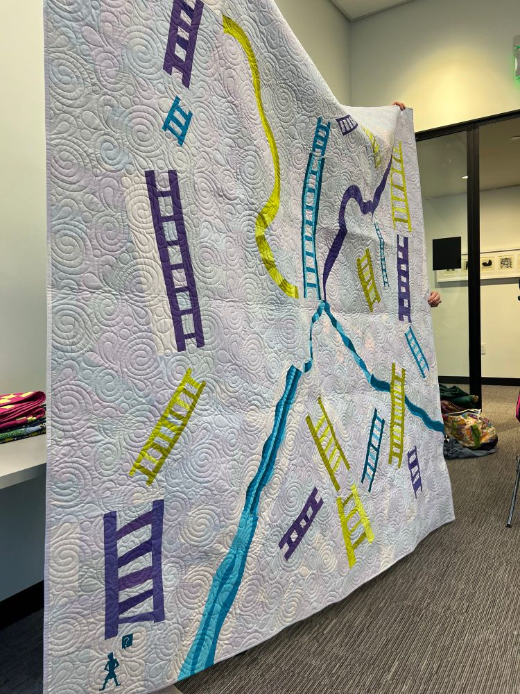 Pale gray & whites background modern quilt with ladders and slides in purple teal and yellow-greens  held aloft by a couple of people not visible except a few fingers. 