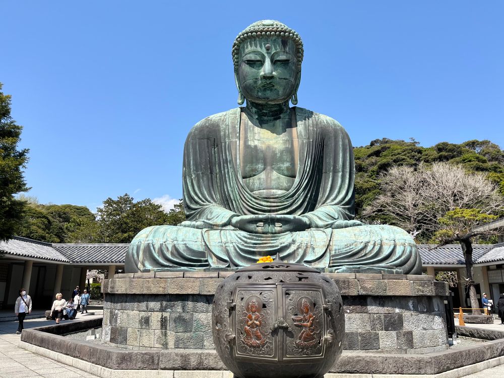 This photo shows the Great Buddha of Kamakura (Kamakura Daibutsu), a famous monumental outdoor bronze statue of Amida Buddha located in Kamakura, Japan. The statue is seated in a meditative posture, with its hands forming the Dhyana Mudra, a common gesture in Buddhist art that represents meditation and balance.

The Buddha sits atop a large stone base, and in the foreground is a decorative bronze incense burner adorned with detailed carvings of Buddhist deities. Surrounding the statue are traditional Japanese buildings and lush trees, with a clear blue sky overhead, giving the scene a calm and serene atmosphere.