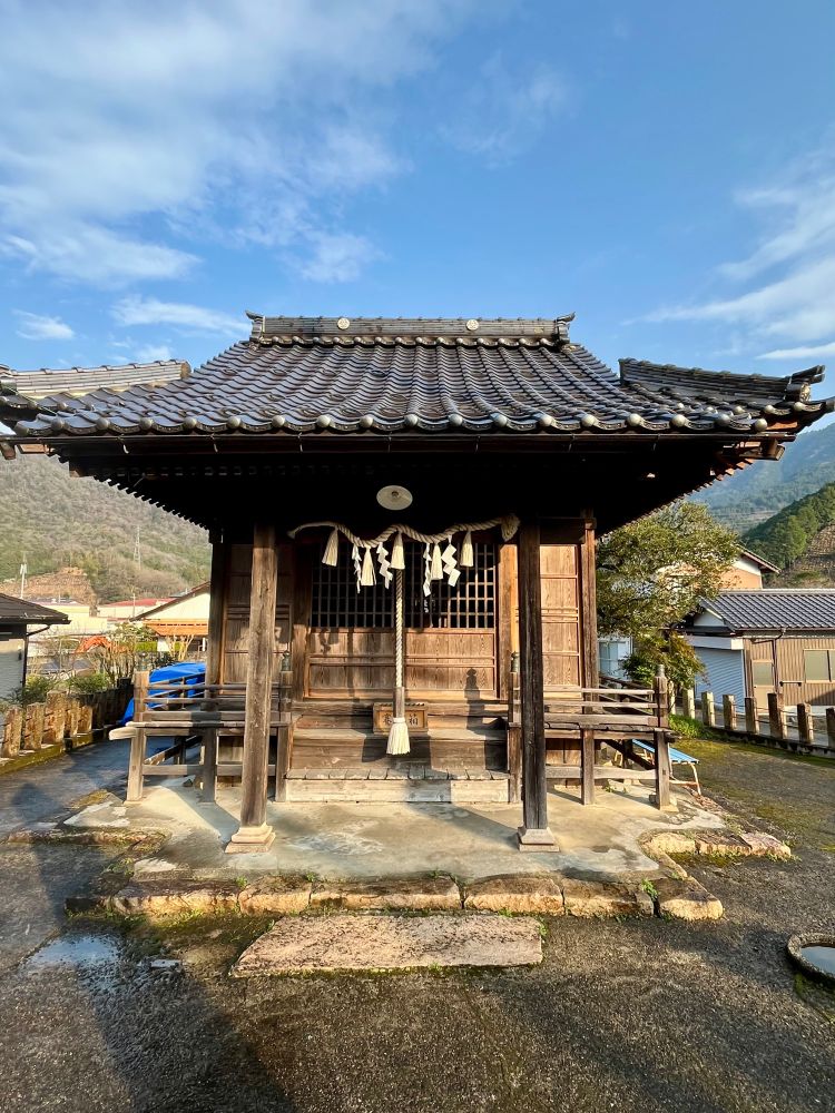The photo shows a small traditional Japanese shrine (Shinto or Buddhist) with a wooden structure and a tiled roof. The architecture features curved eaves, wooden pillars, and intricate craftsmanship. The front of the shrine has a shimenawa (sacred rope) with shide (zigzag-shaped paper strips), which are commonly found in Shinto shrines to signify a sacred space. A large bell with a thick rope hangs at the entrance, likely used for worshippers to call upon the deity before praying.

The setting is in a rural or semi-rural area, surrounded by hills and greenery in the background. The shrine stands on a slightly elevated concrete foundation with stone steps leading up to it. Wooden benches are placed on either side of the entrance. There are a few modern elements such as houses, utility poles, and a blue tarp in the background, indicating the shrine is located within a small village or town. The weather is clear and sunny, casting long shadows, suggesting it was taken in the morning or late afternoon.

Alt text by ChatGPT