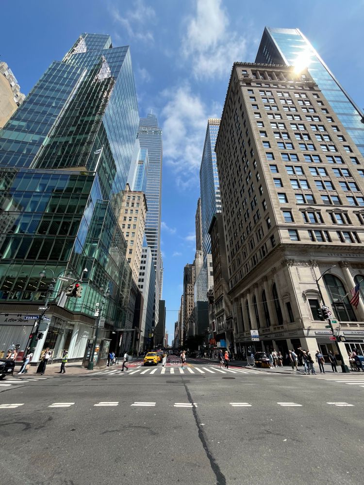 This is a classic Manhattan street scene showcasing the dramatic urban canyon effect created by New York’s towering skyscrapers. The photo captures the beautiful contrast between the city’s architectural eras - you can see older pre-war buildings with their classic brick and stone facades alongside sleek modern glass towers that reflect the brilliant blue sky.

The perspective from street level really emphasizes the scale and density of Manhattan’s built environment. The wide avenue stretches into the distance, flanked by these architectural giants that seem to reach endlessly upward. You can see pedestrians crossing at the crosswalk, giving a sense of the human scale against these massive structures.

The lighting is particularly striking - the sun appears to be hitting one of the glass buildings, creating that bright glare, while the deep blue sky with white clouds provides a stunning backdrop. The street appears to be relatively quiet for Manhattan, which might suggest this was taken on a weekend or during a less busy time of day.

This type of view is quintessentially New York - the interplay of old and new architecture, the urban canyon effect, and the way the city’s grid system creates these dramatic sight lines down the avenues. It’s the kind of scene that captures why Manhattan’s streetscape is so iconic and instantly recognizable around the world.​​​​​​​​​​​​​​​​