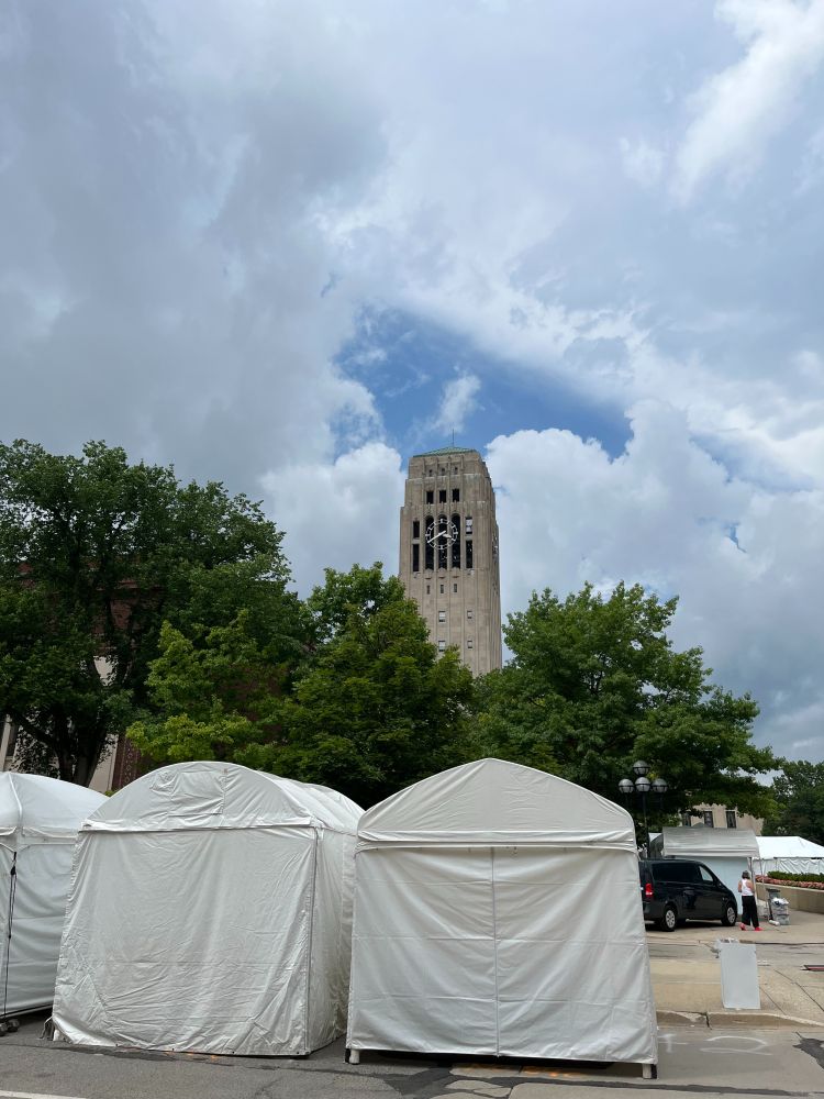 This photo captures a scene in downtown Ann Arbor, Michigan, featuring the iconic Burton Memorial Tower in the background. The tower, part of the University of Michigan campus, stands tall behind a group of white vendor tents that are set up for the Ann Arbor Art Fair.

In the foreground are several closed, peaked white tents arranged in a row, set up on a blocked-off street. Trees with lush green foliage partially obscure the base of the tower, and a mostly cloudy sky with a patch of blue peeking through fills the top of the image. To the right, a person stands near a black van, suggesting early setup or preparation for the event.
