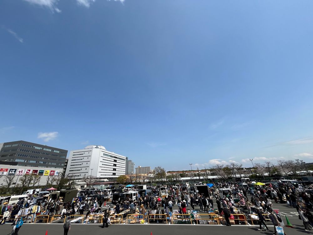 This photo shows a bustling outdoor flea market or bazaar, likely held in a large parking lot. The scene is filled with numerous stalls and tables, where vendors have laid out various goods — possibly secondhand clothes, antiques, or collectibles. The market is busy, with many people browsing and shopping.

In the background, there are several modern buildings, including a white multi-story building and a dark gray office complex with Japanese signage. The signs suggest that this market is taking place in Japan. The sky is clear and bright blue with minimal cloud cover, indicating a beautiful, sunny day. 