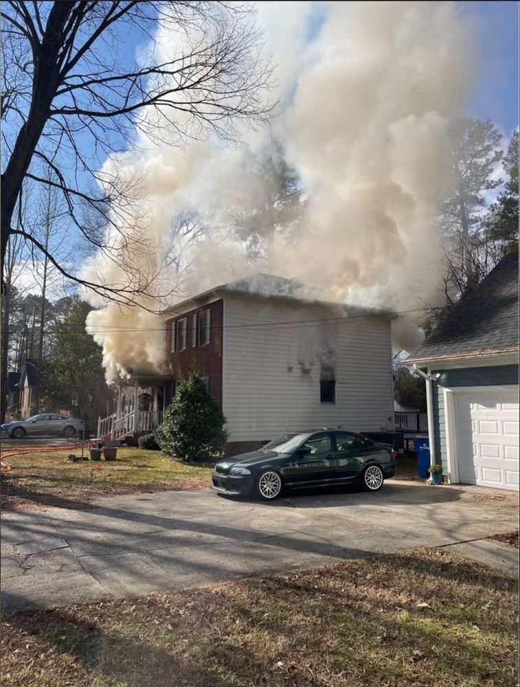 a photo of a a dark green 1999 bmw 3 series in the driveway of a suburban home in front of the garage. in the background, the neighbor's house is clearly on fire as there is smoke billowing from the door and windows.