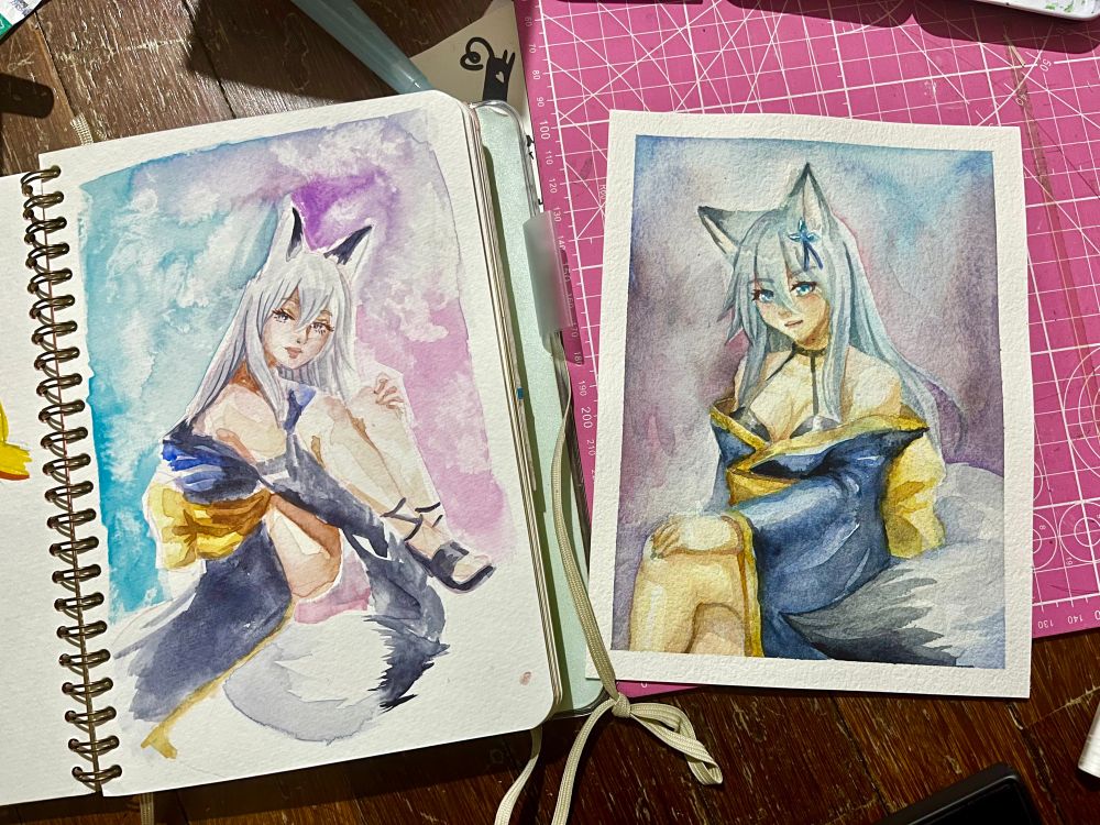 A picture of a sketchbook next to a loose sheet painting on top of a pink cutting mat. The sketchbook has an illustration of my original character Sakura, a white fox spirit in a loose kimono sitting with her knees bent while holding her legs. The loose sheet painting is also an illustration of my original character Sakura done in a similar pose facing the other direction, except it is 3/4 body instead of full body. The two paintings were done 7 years apart. Picture was taken in a way to showcase the improvement in watercolour technique