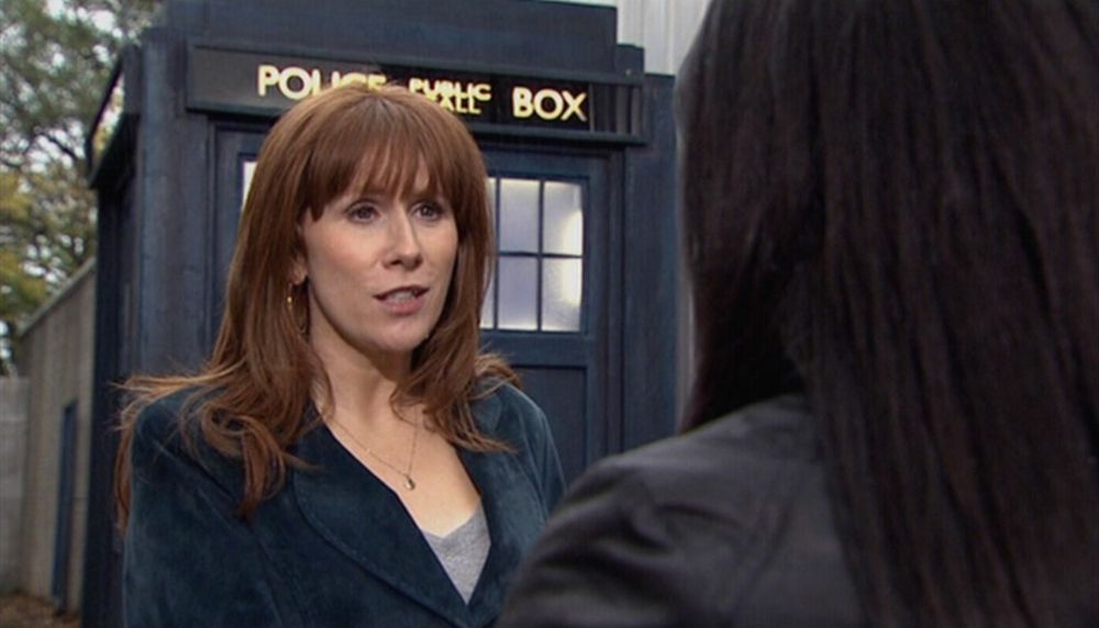 Catherine Tate as Donna Noble in Doctor Who.
She's a middle-aged white woman with long red hair, wearing a navy blazer over a pale grey low-cut top. She looks like she's mid-conversation with someone, while standing in front of the TARDIS (an old-style British police box (like a large telephone booth)).