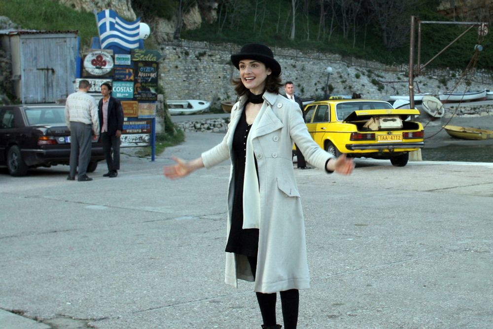 Rachel Weisz as Penelope Stamp in The Brothers Bloom.
She's a white woman wearing black tights, a black dress, and a pale khaki trench coat. Her shoulder-length hair is under a broad-brimmed black hat as she stands, smiling eagerly, arms extended to the sides, at a dockyard.