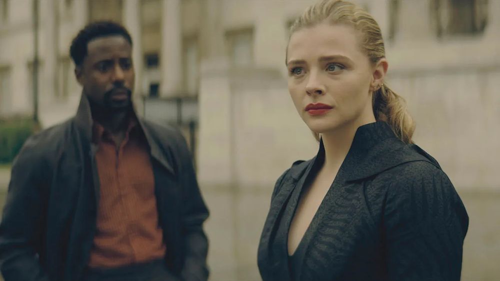 A Black man with short hair and a goatee (Gary Carr) stands in a red shirt and black jacket, looking at a white woman (Chloe Grace Moretz) who is looking off into the distance, in a very high-tech looking black jacket with deep v-neck, her blonde hair pulled back.

They are standing on an urban street surrounded by white stone buildings.
