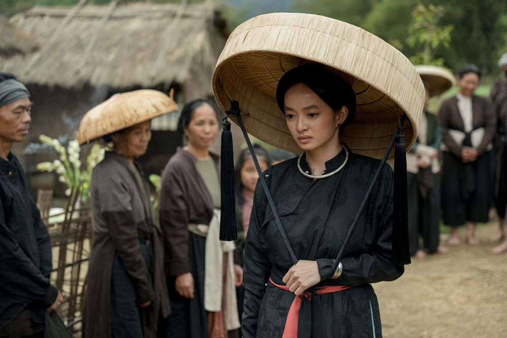 Kaity Nguyen as Linh in The Last Wife
A young Vietnamese woman wearing historical peasant clothing (late 1800s??? She's got a long-sleeved black blouse and a black skirt tied with a red belt. She's wearing and carrying a large woven hat shaped like a large flat-bottomed basket with about 8 inches of woven sides; it is being held on her head by the strap she's holding in her hand, down by her waist. She looks downtrodden and not happy as she walks past a long line of fellow villagers.