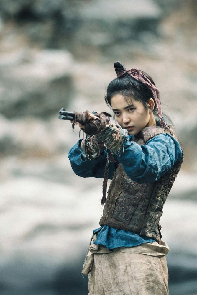 Kim Tae-ri as Go Ae-shin in Mr Sunshine.
A young Korean woman, standing, aiming with a sniper rifle.
She's wearing historical clothes from the early 1900s in Korea... scrappy, dirty warrior clothes - wide-legged woven pants, a long-sleeved shirt of some kind under a padded vest (likely for warmth as much as anything else, and wraps of rags around her hands and the muzzle of her gun, to dampen the sound.
Her hair is up in a men's style topknot, tied with another strip of rag.
