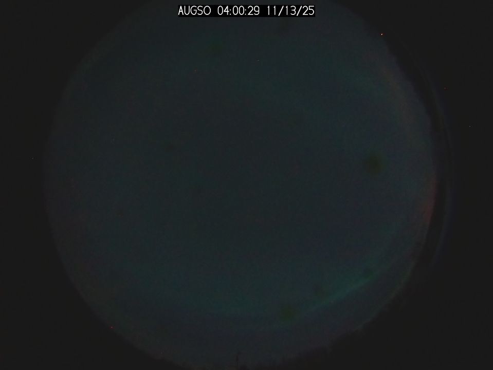 A circular photo of the whole night sky, showing a faint wash of green across most of the sky, bolder streaks arc from right to left across the bottom curve and the top curve. There's also a hint of red along the outer edge., it's more visible on the right hand side.
