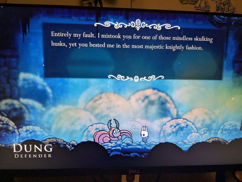 An image from Hollow Knight- the red rotund dung beetle perched in the poopy earth next to the tiny hollow Knight. The caption says- " entirely my fault. I mistook you for one of those mindless skulking husks, yet you bested me in the most majestic knghtly fashion."
