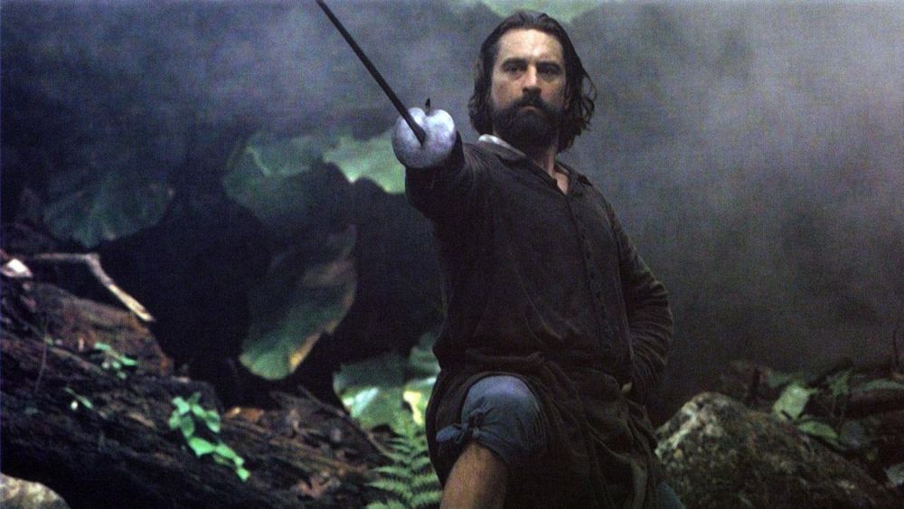 A still from THE MISSION (1986). Robert De Niro, with a black beard and swept-back dark hair, stands in a haze of smoke in a jungle scene. He is dressed in a black shirt, and is holding a rapier out in front of him in his right hand. His left is on his hip, in a fencer's fighting stance. He looks unsmilingly along the length of his sword to whatever faces him.