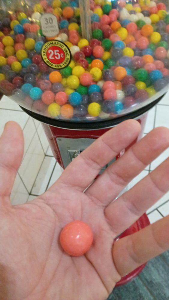 A picture of a left hand holding a gumball, in front of a standalone coin-op gumball machine. The color of the gumball is uncomfortably close to the color of the hand's skin.