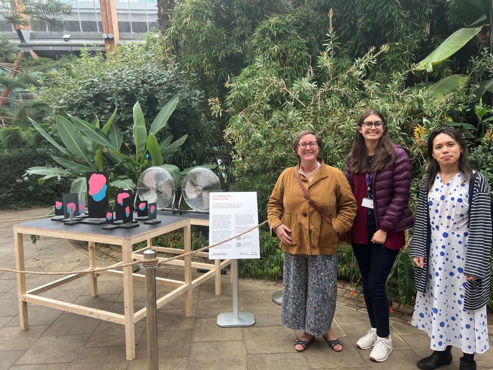 Three members of the RiboCode team stand beside an exhibit in an indoor garden.