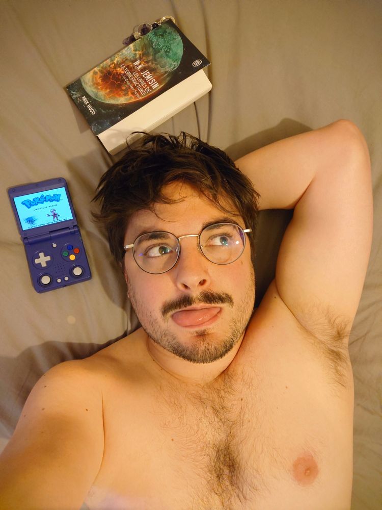 Selfie torse nu sur mon lit avec ma console anbernic RG 34XXSP en train de jouer à Pokémon version bleue et le tome 1 des livres de la terre fracturée