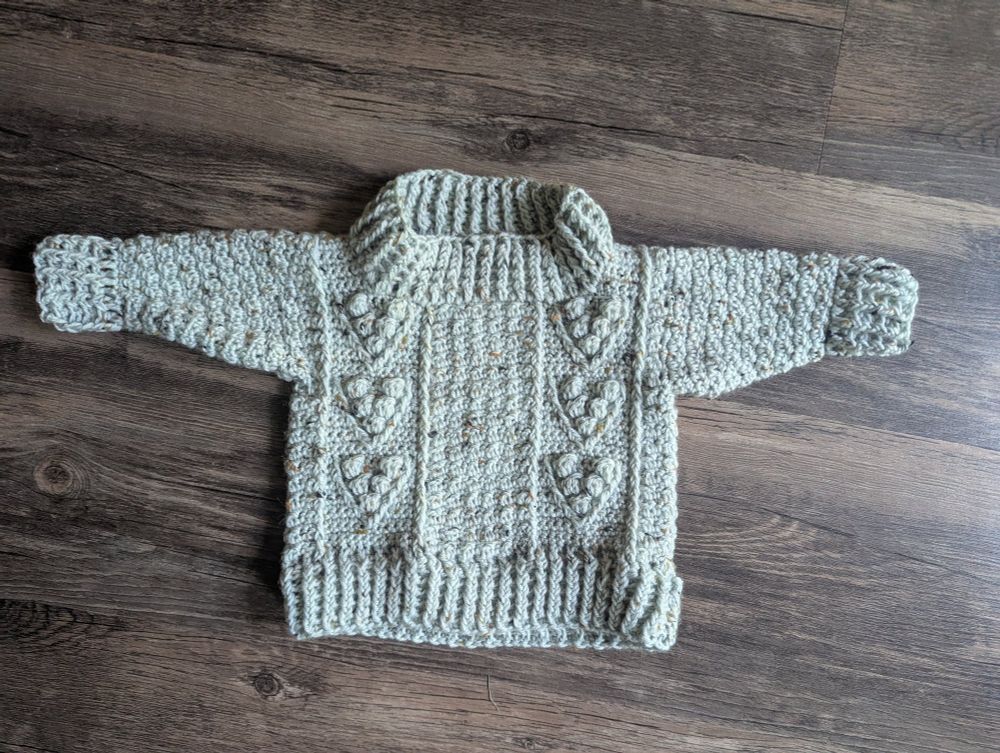 A crocheted child's sweater with detailing that looks like batches of berries. The yarn is a off white tweed. It's against a wooden looking background 