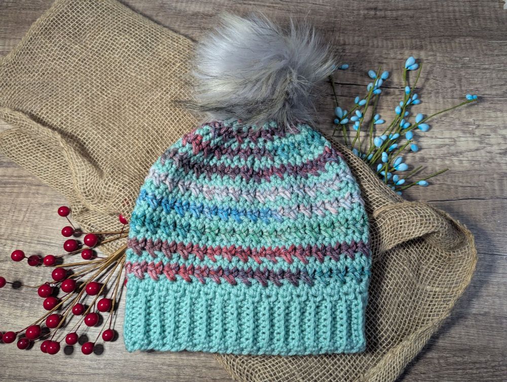 A crocheted hat that is mint and variegated striped. The hat has a purpleish gray faux fur pompom on it. 