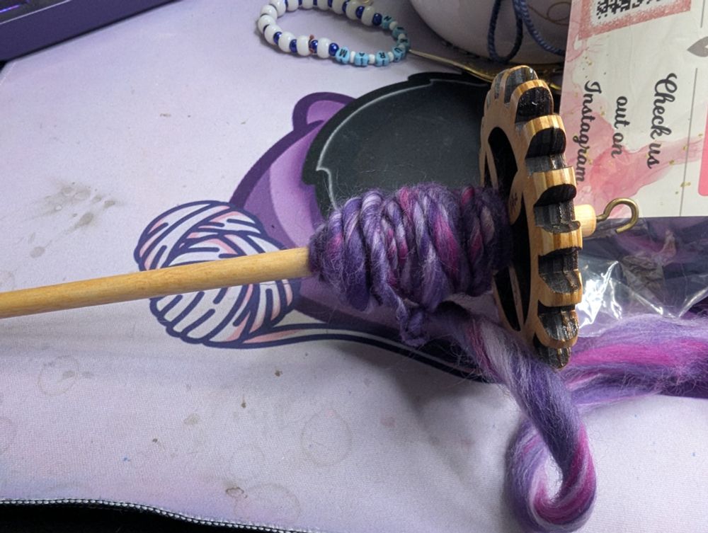 A picture of a drop spindle with yarn being spun on it. The fiber is varying shades of purple. 