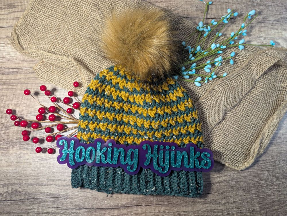 A crocheted hat. The hat uses dark green and gold tweed yarns. On top of the hat is a golden brown faux fur pompom 