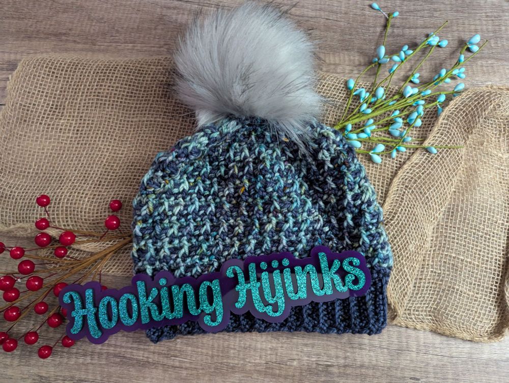 A picture of a crocheted hat. The item is a dark blue mixed with a variegated blue. The hat has a gray faux fur pompom. 