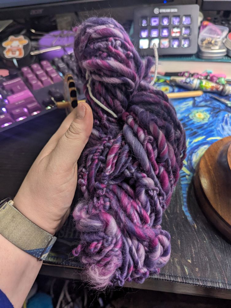 A picture of a hank of hand spun yarn. The yarn is thicker and is in varying shades of purple. 
