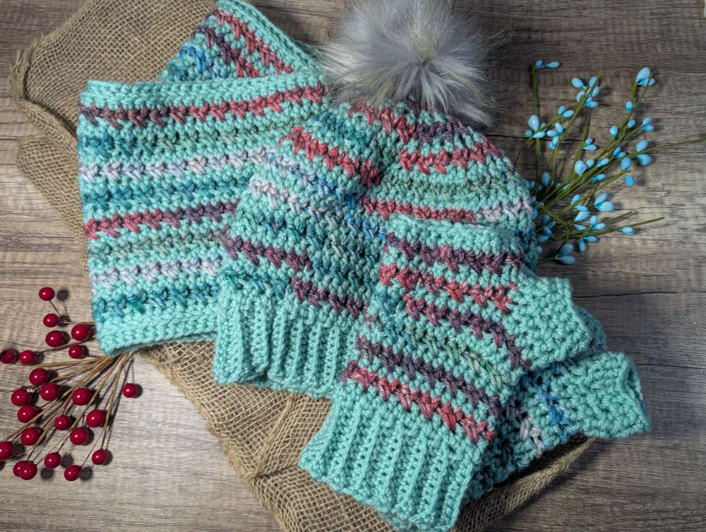 A crocheted winter accessories set. There is a cowl, hat, and fingerless mitts. The items are in a mint green with a variegated matching yarn as stripes. The hat has a purpleish gray faux fur pompom. 