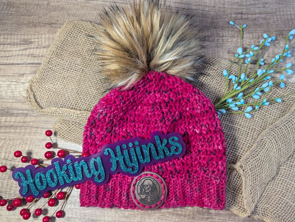 A crocheted hat that is hot pink. The hat has a brown faux fur pompom and a leather tag on the brim that has a skeleton on it. 