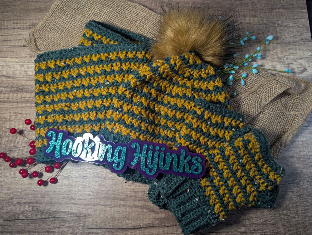 A crocheted winter set. The hat, fingerless mitts, and cowl are striped in a dark green tweed and a golden tweed yarns. The hat has a golden brown faux fur pompom 