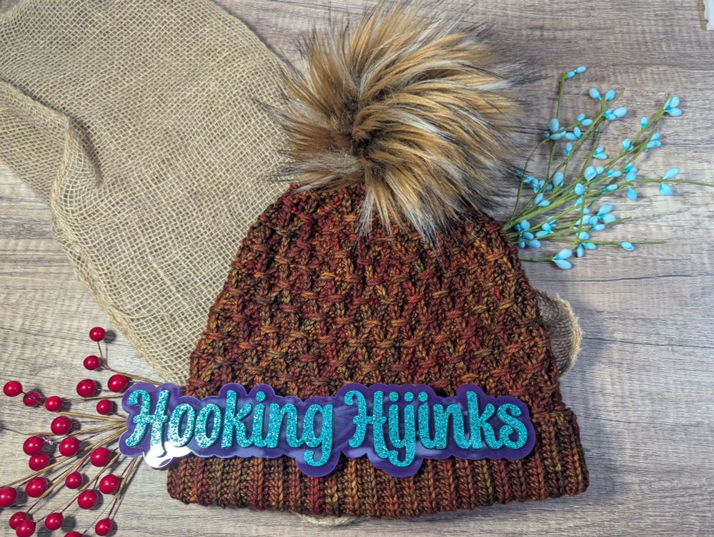 A crocheted hat with an interlacing weave pattern. The yarn used is various shades of orange and there is a brown faux fur pompom on the top. 