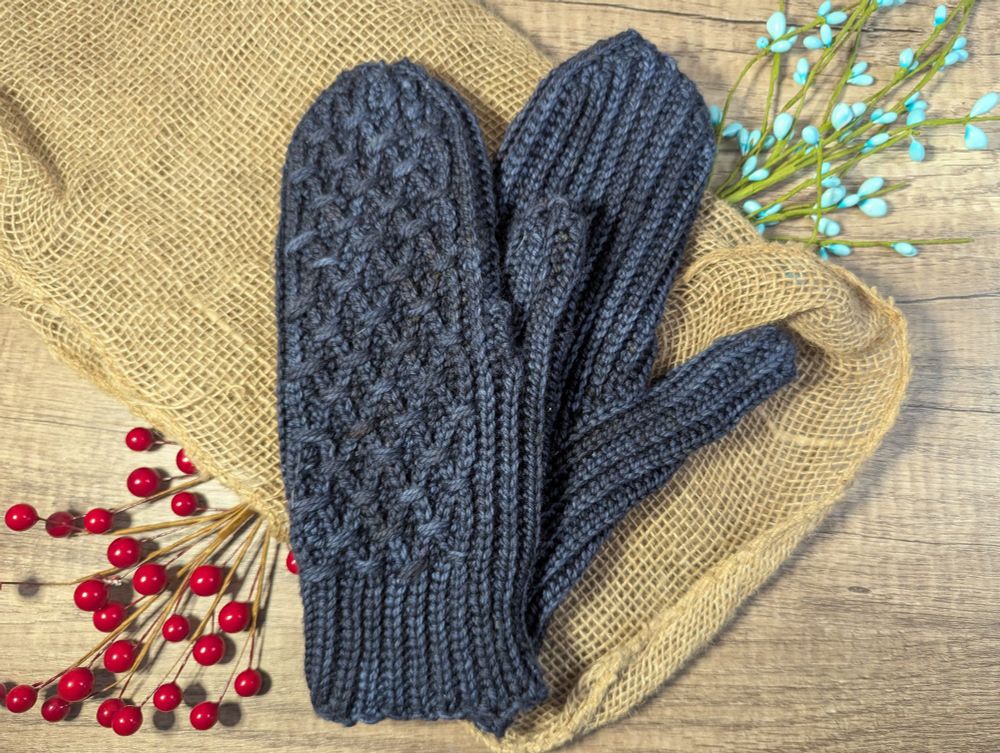 Crocheted dark blue mittens against a brown and floral flat lay background. The back of the mittens has an interlacing pattern of stitches and the item almost looks knitted 