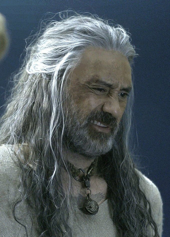 Taika as Ed in the rice-sack onesie and cat bell,  making a face that suggests he's got something caught in the concealed crotch zipper. 
